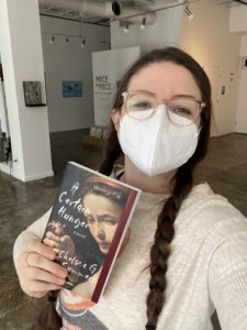 Store manager Emily, masked, holding a copy of A Certain Hunger